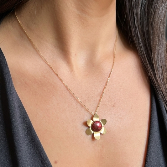 A close-up of a person wearing a delicate gold chain necklace with a flower-shaped pendant featuring a round, dark red center, paired with a dark gray top.