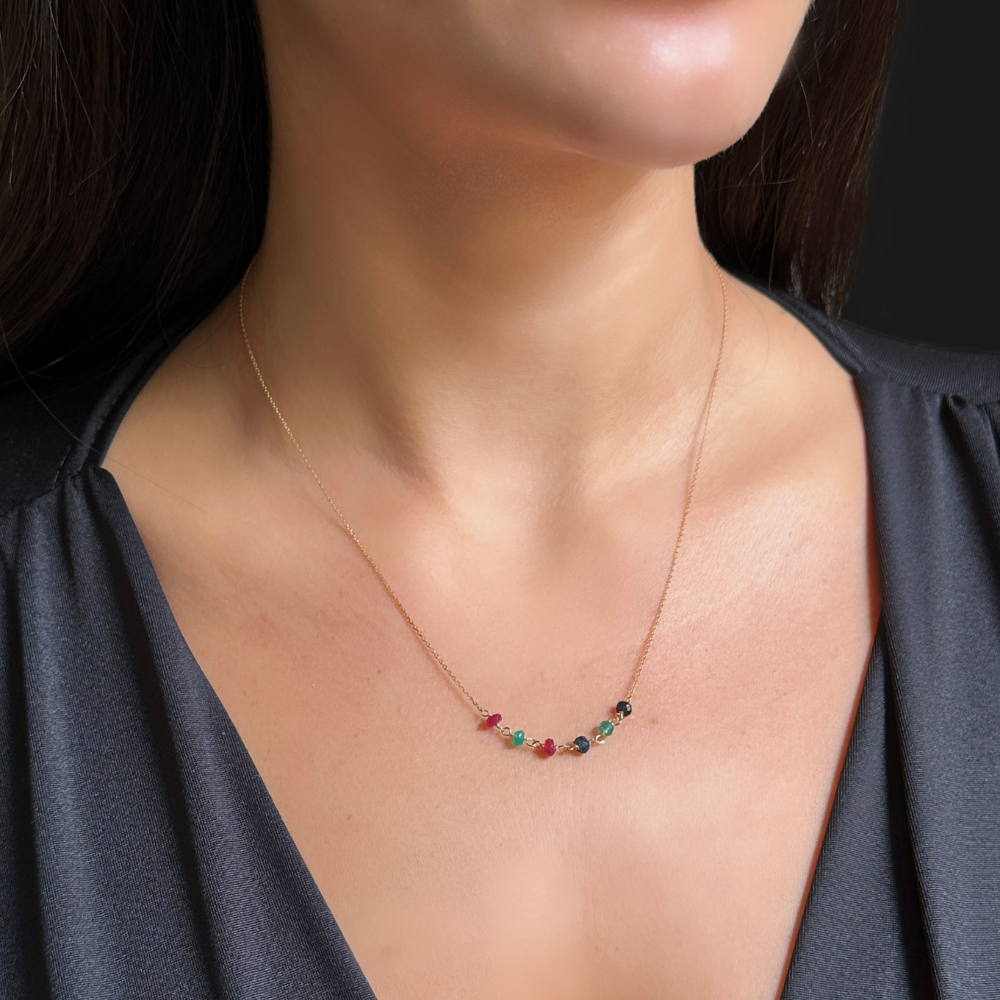 A close-up of a model wearing a delicate gold necklace with a curved row of small, multi-colored gemstone beads at the center, paired with a dark gray V-neck top.