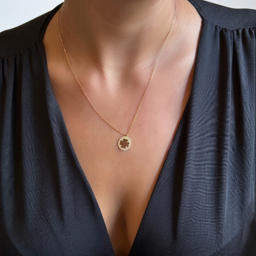 A close-up of a person wearing a delicate gold necklace with a circular pendant featuring a clover-like design, paired with a dark navy blue blouse.