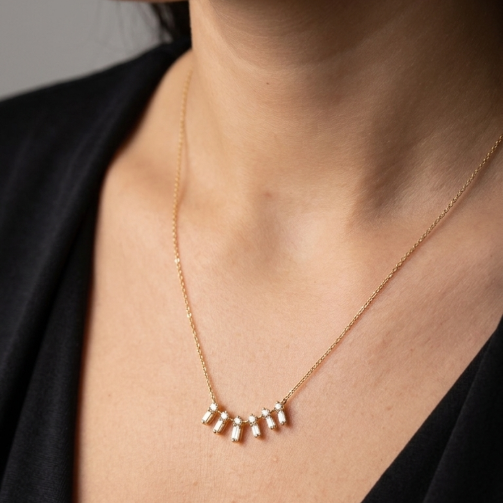 A close-up of a person's neck and chest wearing a delicate gold chain necklace with five small dangling bar charms, styled with a black top. Zircon baguettes – gold necklace