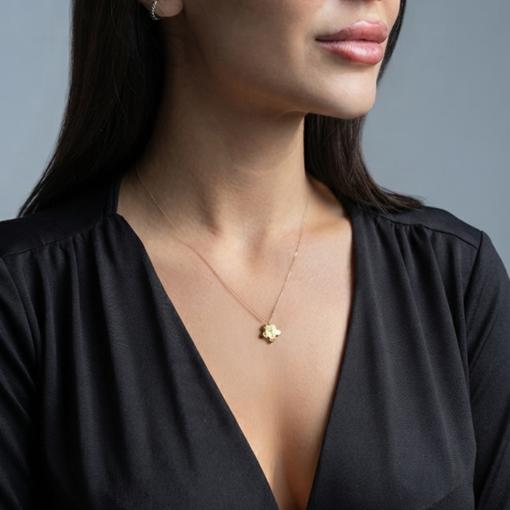 A close-up of a woman wearing a delicate gold necklace with a small flower-shaped pendant, paired with a black V-neck top against a solid gray background. Yellow gold flower necklace