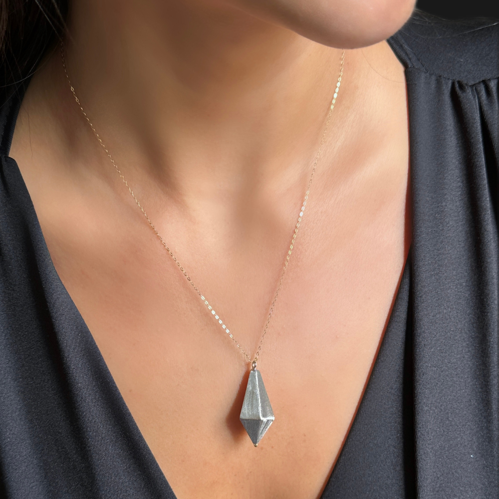 A close-up of a person wearing a delicate gold chain necklace with a faceted silver geometric pendant, styled with a dark gray V-neck top.