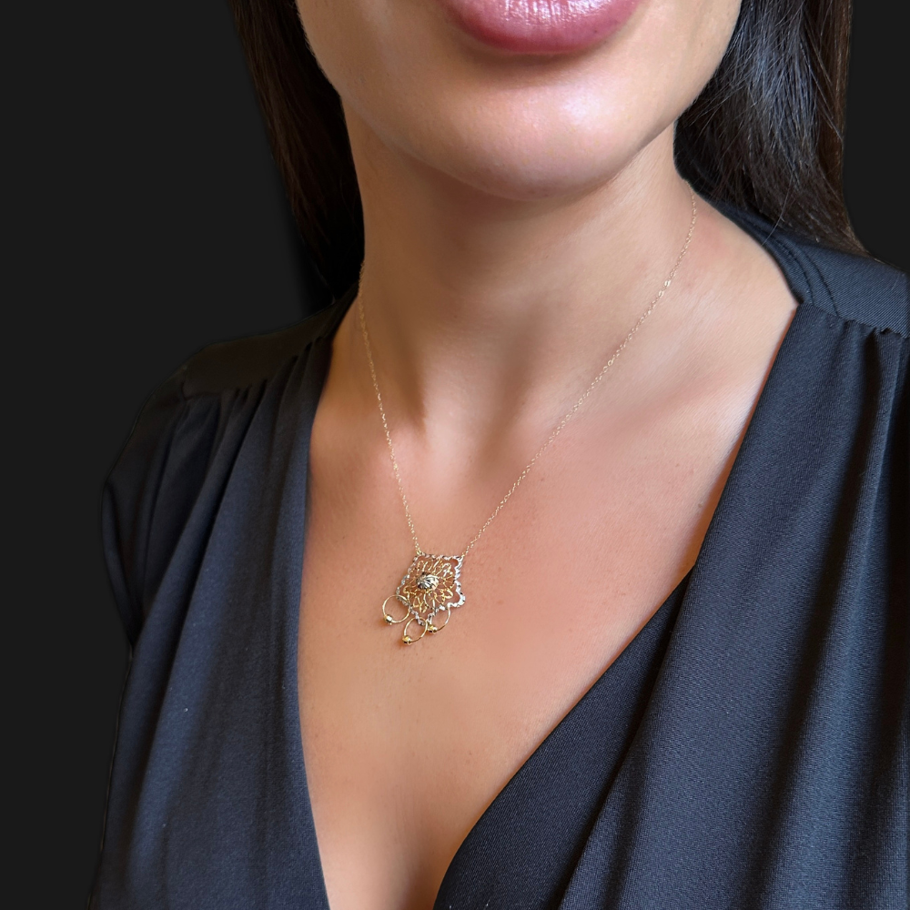 A close-up of a woman wearing a delicate gold necklace with a geometric, flower-like pendant featuring a central clear stone. She is wearing a black V-neck top against a dark background.