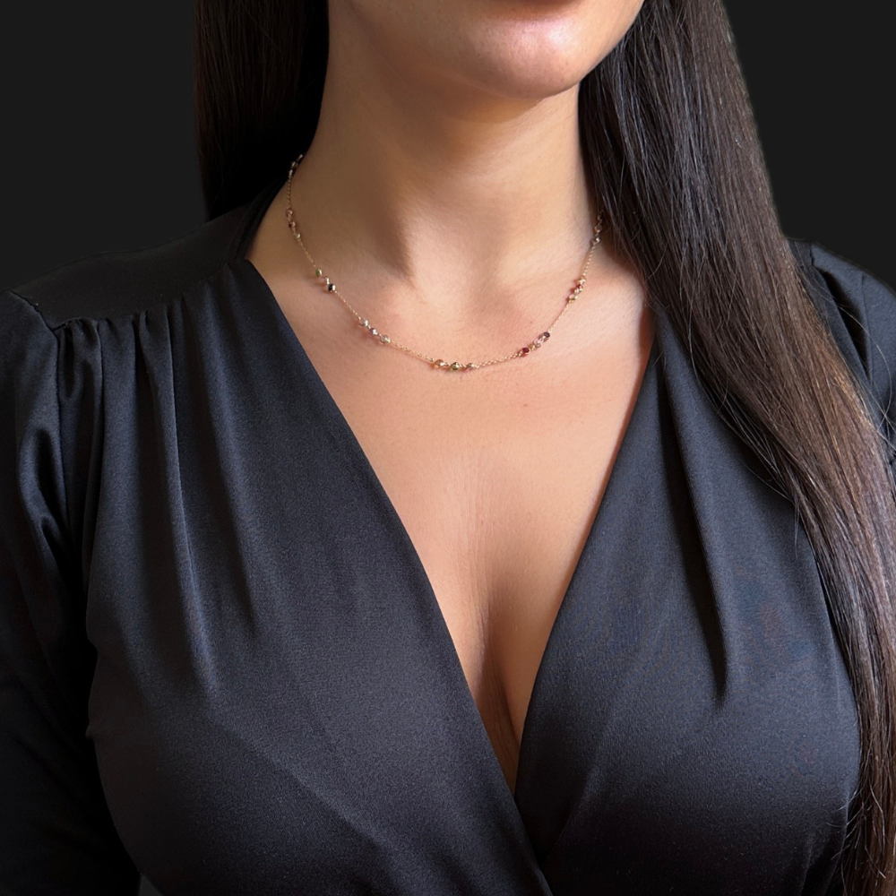 A close-up of a woman wearing a delicate gold necklace with small heart-shaped beads, paired with a black satin V-neck top against a dark background.