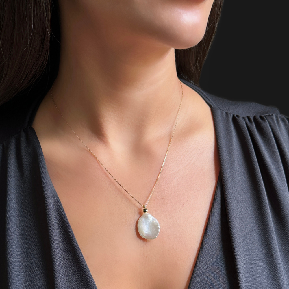 A close-up of a woman wearing a delicate gold chain necklace with a teardrop-shaped white pearl pendant, paired with a dark gray V-neck top.