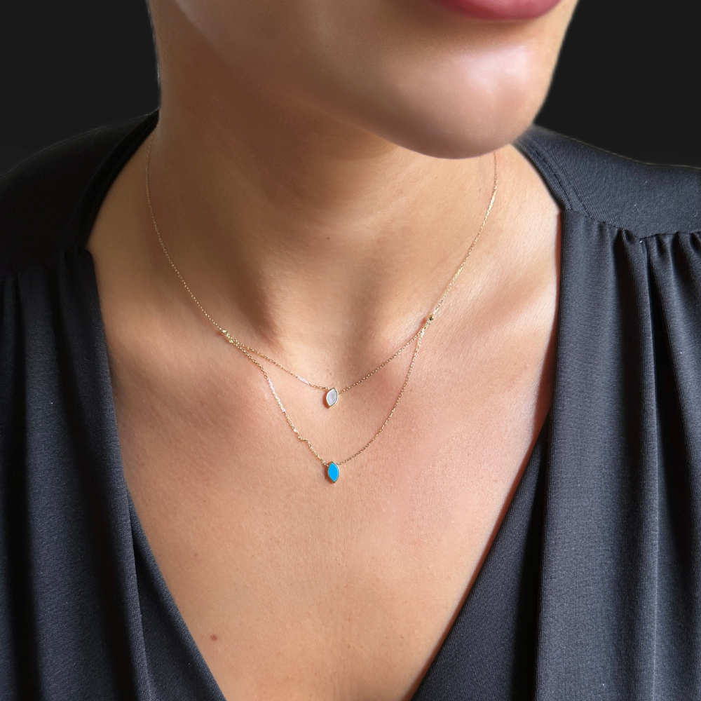 A close-up of a model wearing a delicate layered gold necklace with a small white teardrop pendant and a longer chain featuring a small bright blue teardrop pendant, paired with a dark gray V-neck top.