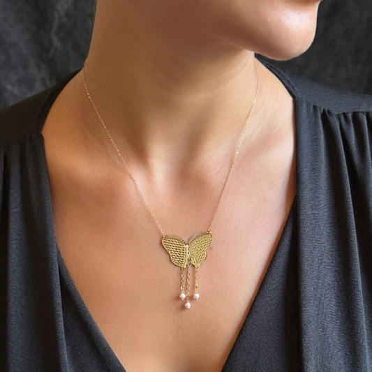 A close-up of a model wearing a delicate gold chain necklace with a butterfly-shaped pendant and dangling pearl beads, styled with a dark gray V-neck top.
