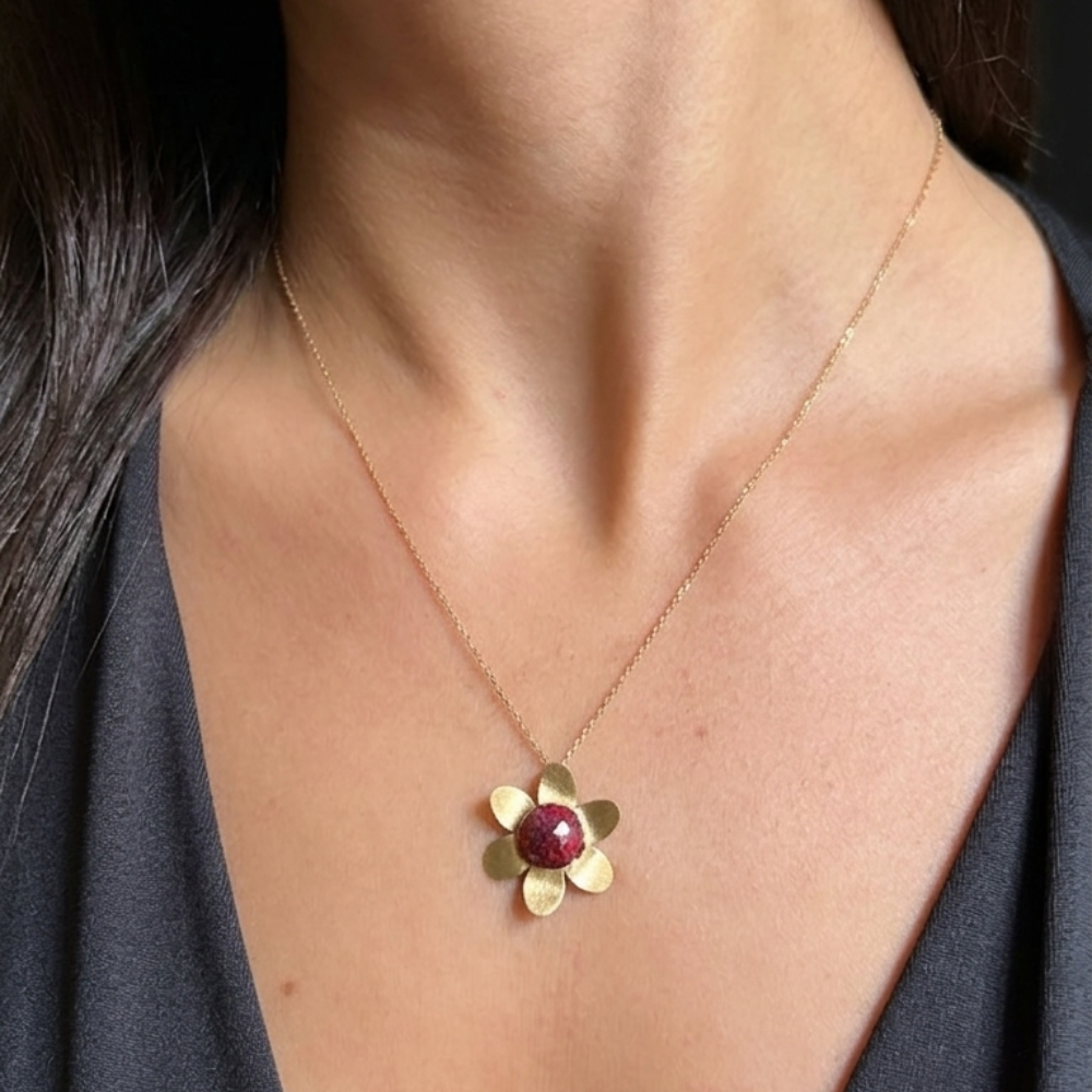 A close-up of a person wearing a delicate gold flower pendant necklace with a faceted red stone center, paired with a dark gray top.