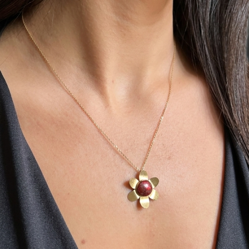 A close-up of a person wearing a delicate gold chain necklace with a flower-shaped pendant featuring a round, dark red center, paired with a dark gray top.