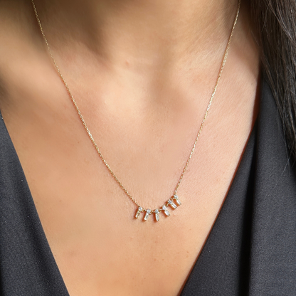 A close-up of a person's neck and chest wearing a delicate gold chain necklace with a small letter pendant necklace spelling 'FAMILY' in clear stones. Zircon baguettes – gold necklace