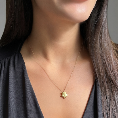A close-up of a model wearing a delicate gold chain necklace with a small gold flower or clover pendant, paired with a black V-neck top. Yellow gold flower necklace
