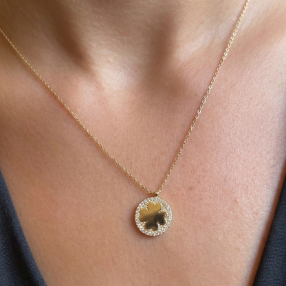 A close-up of a gold necklace with a clover-shaped pendant, worn on a person's neck over a dark gray top.