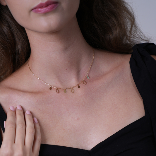 A close-up of a woman wearing a delicate gold chain necklace with small dangling oval charms, paired with a black off-the-shoulder top.