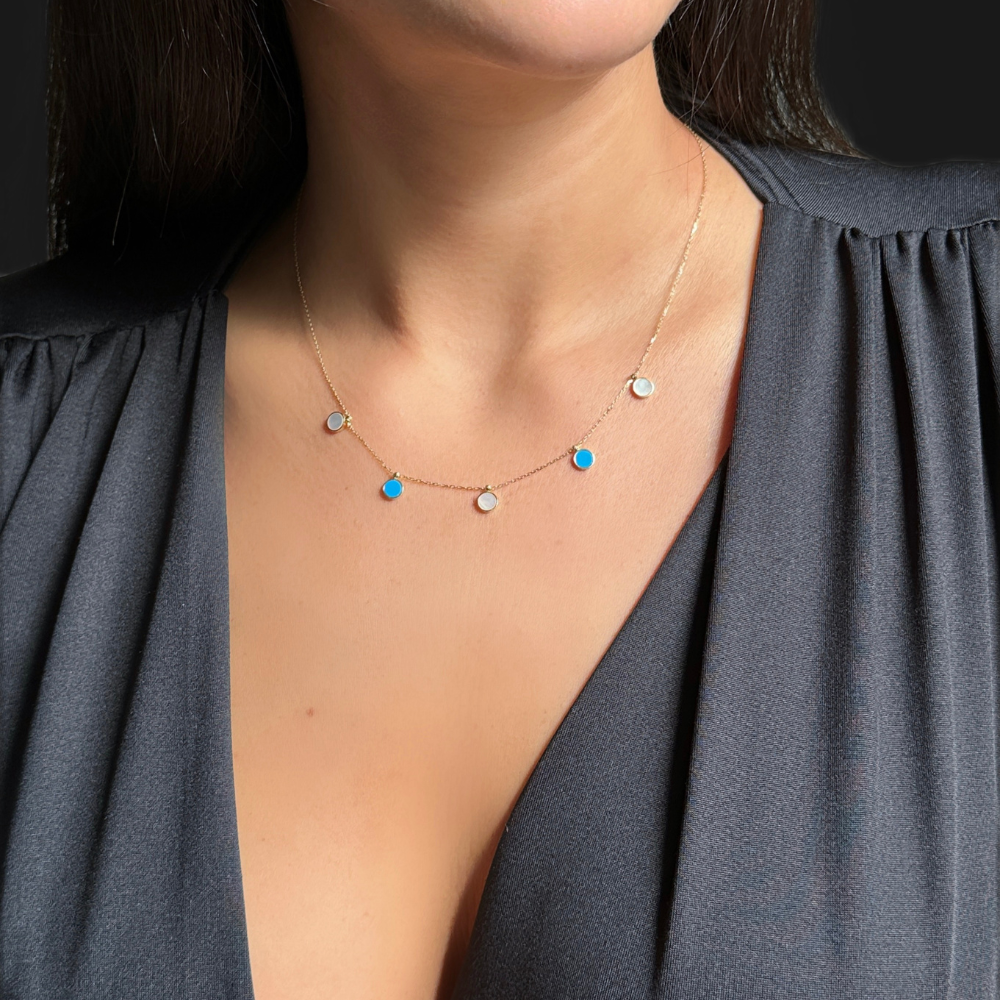 A close-up of a woman wearing a delicate gold chain necklace with small mother of pearl and blue circle charms, paired with a dark gray V-neck top.