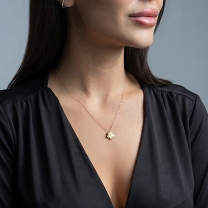 A close-up of a woman wearing a delicate gold necklace with a small flower-shaped pendant, paired with a black V-neck top against a solid gray background. Yellow gold flower necklace