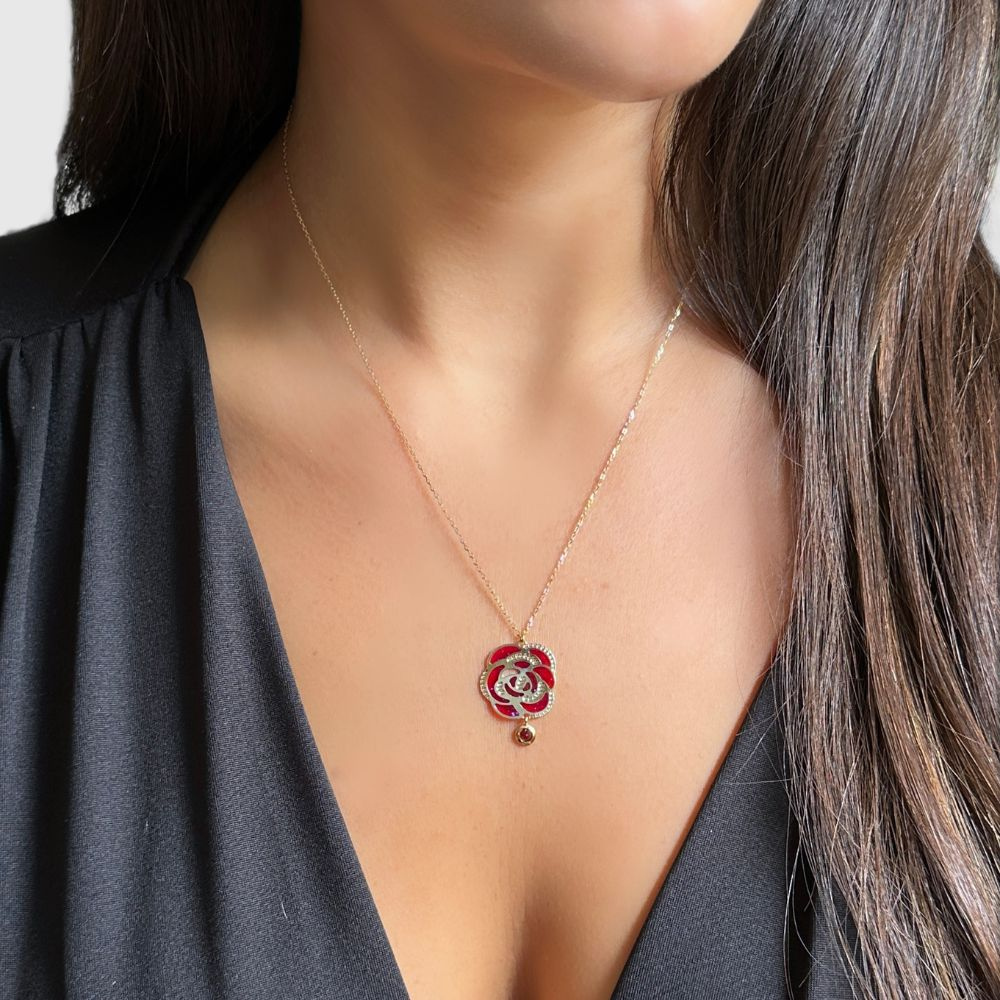 A close-up of a woman wearing a delicate rose-shaped pendant necklace with red and white enamel detailing and a small dangling charm, styled with a black V-neck top. Red rose flower shaped stained pendant - 18k gold necklace