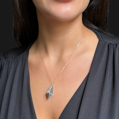 A close-up of a woman wearing a delicate gold chain necklace with a faceted geometric pendant, paired with a dark gray V-neck top.