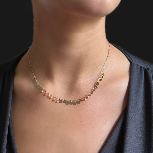 A delicate gold chain necklace with small round coin-like charms, worn on a model's neck over a dark gray top. Circles charm gold – small necklace